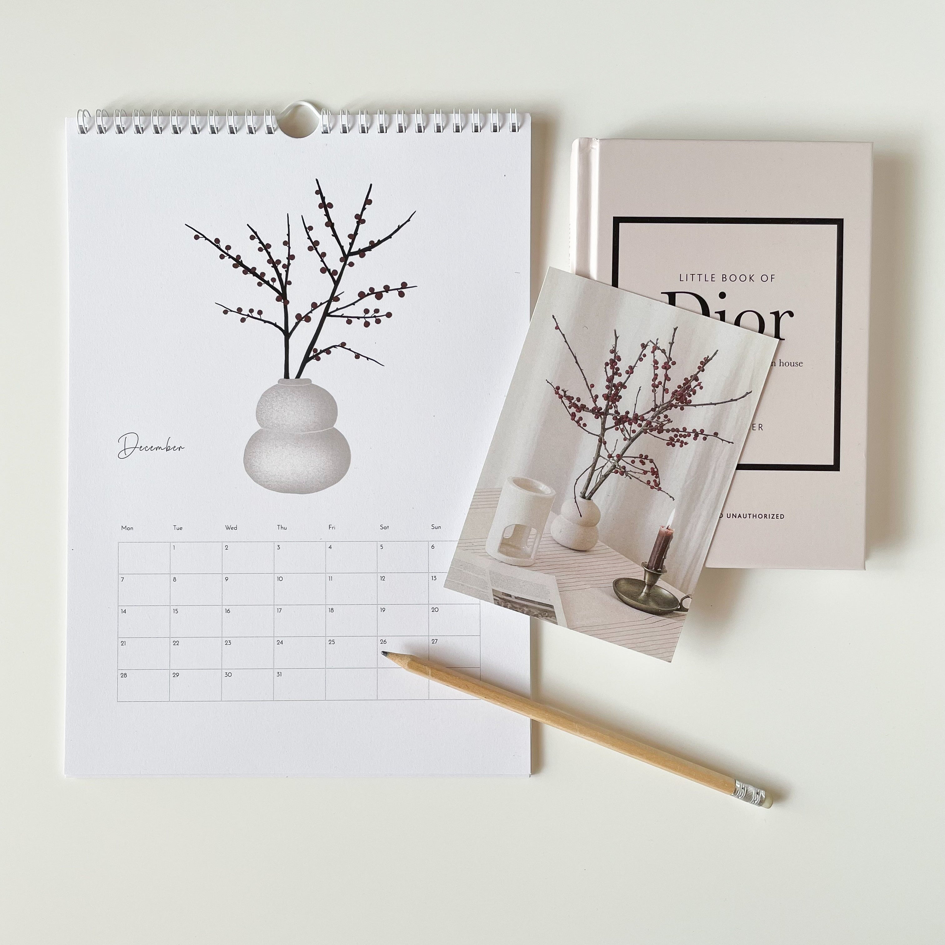 Calendar with a decorative plant illustration, a book titled 'Little Book of Dior', and a pencil on a light background.